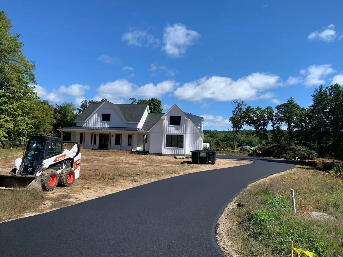 New asphalt driveway curving up to a white farmhouse with a track loader on the yard