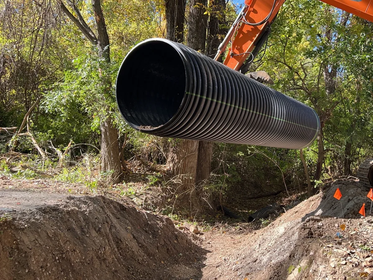 Excavator lowering a large drainage culvert pipe into a cut