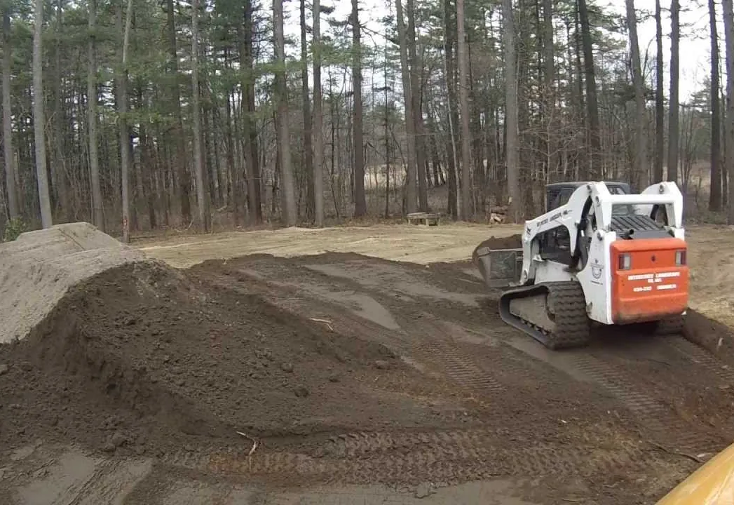 Track loader spreading and grading a sandy dirt pad in a stand of pines