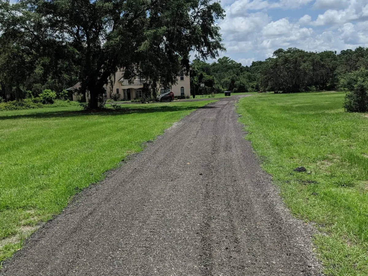 Gravel lane running across a mowed field toward a house under big oaks