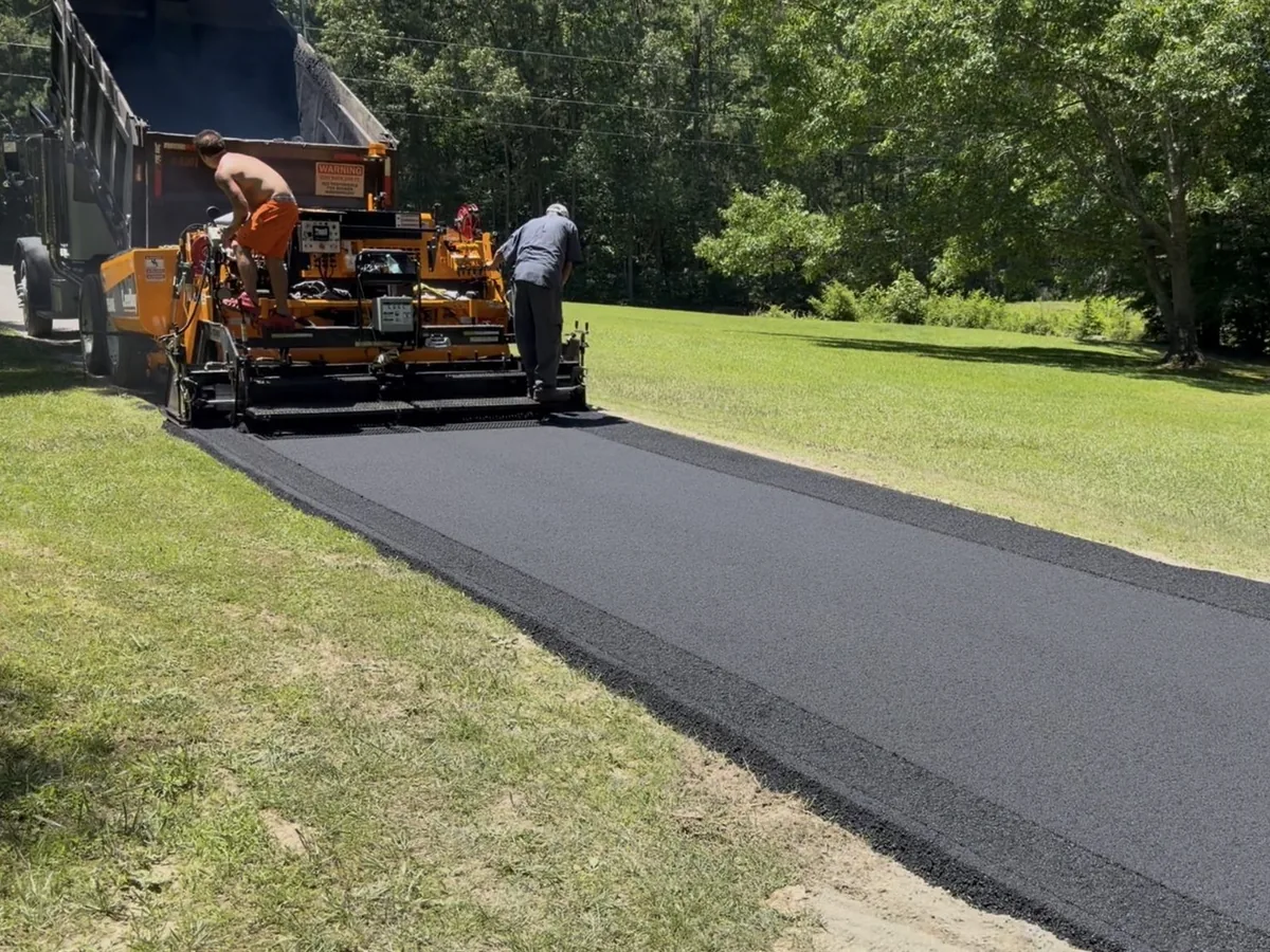 Paver laying a wide mat of fresh asphalt along a grassy shoulder