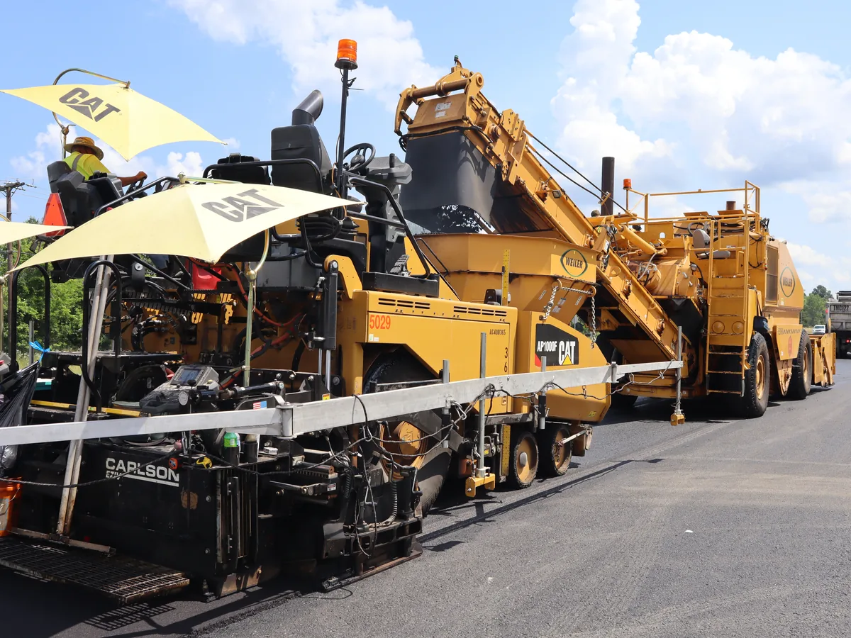 Paver laying a new asphalt surface down a road with a haul truck ahead of it